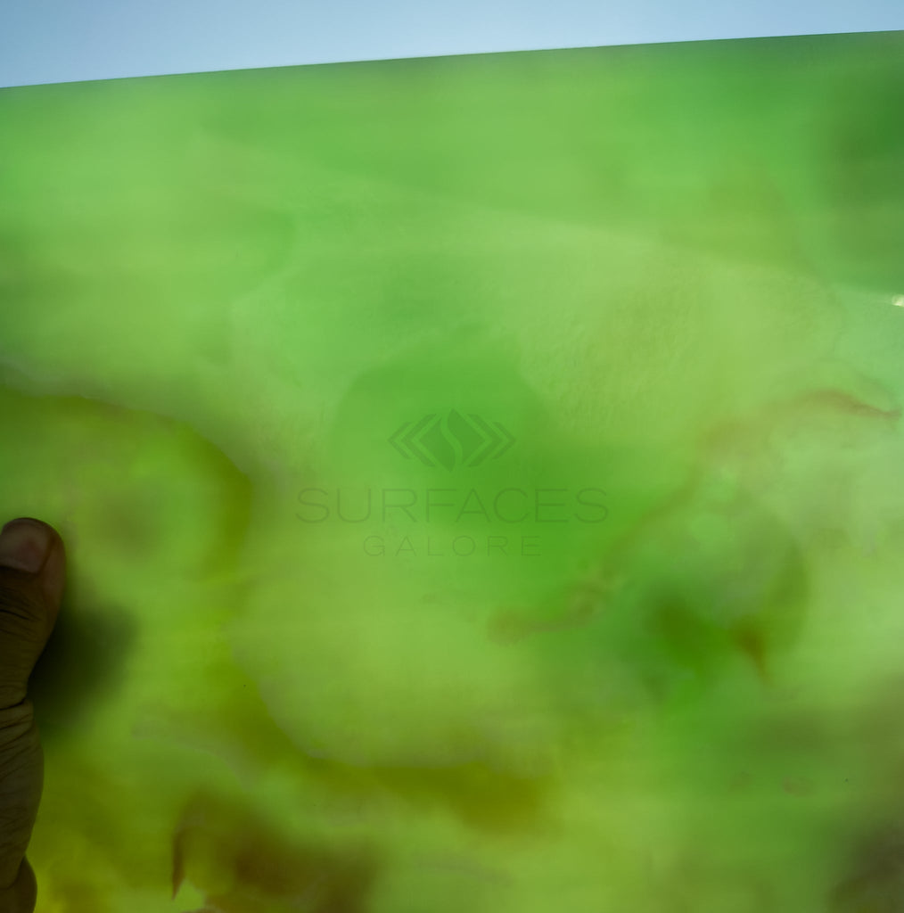 A hand holds a translucent sheet of Green Onyx Premium Marble 12X12 Polished with light passing through. The text "SurfacesGalore" is visible, highlighting their luxury tile collection.