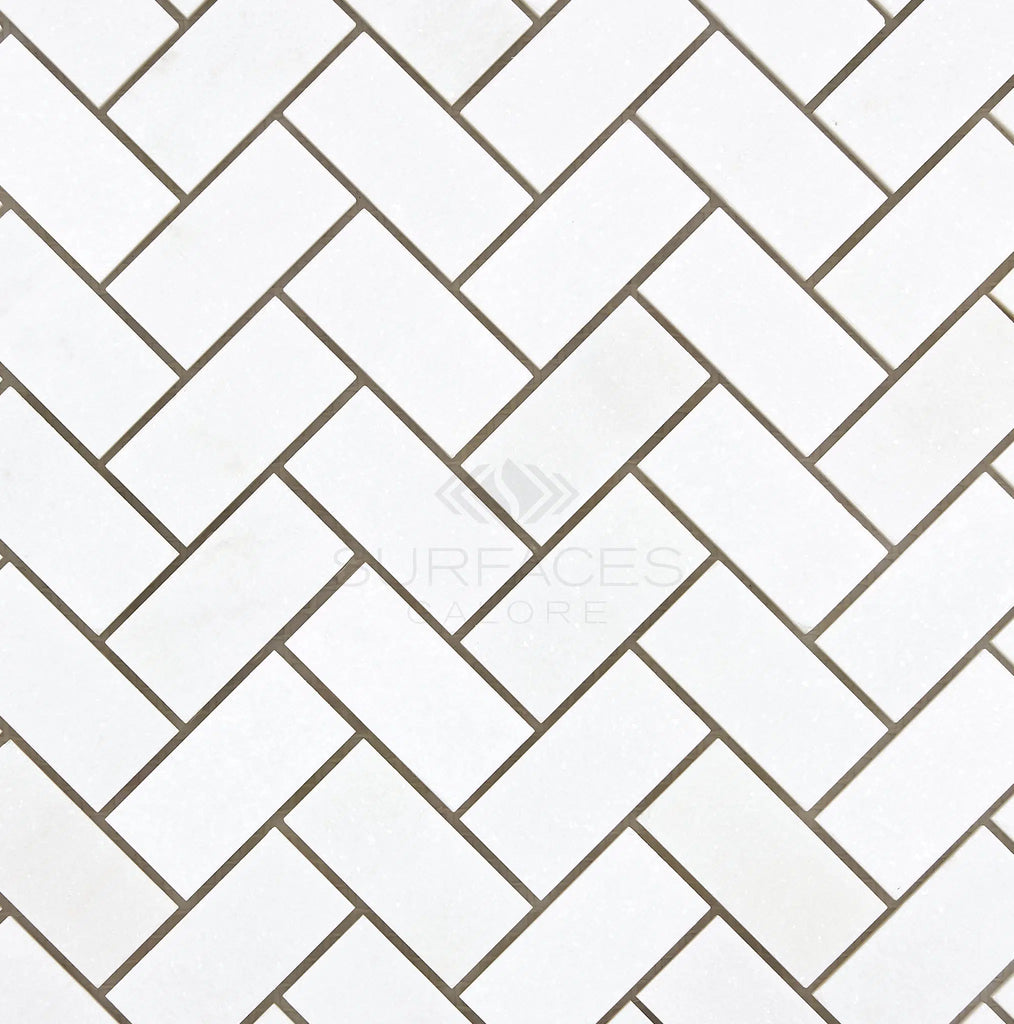 Thassos White (Greek) 1X2 Herringbone Mosaic Marble Polished-Honed tiles with grey grout lines create a chevron pattern, featuring the SurfacesGalore logo at the center.