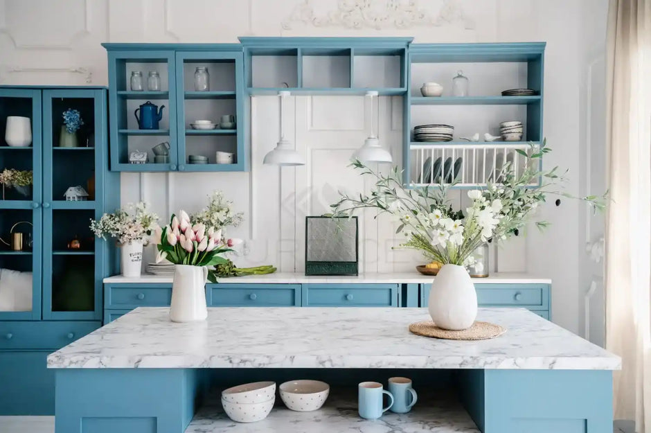 A bright, blue and white kitchen with an island, open shelving, flowers, and dishes neatly arranged.