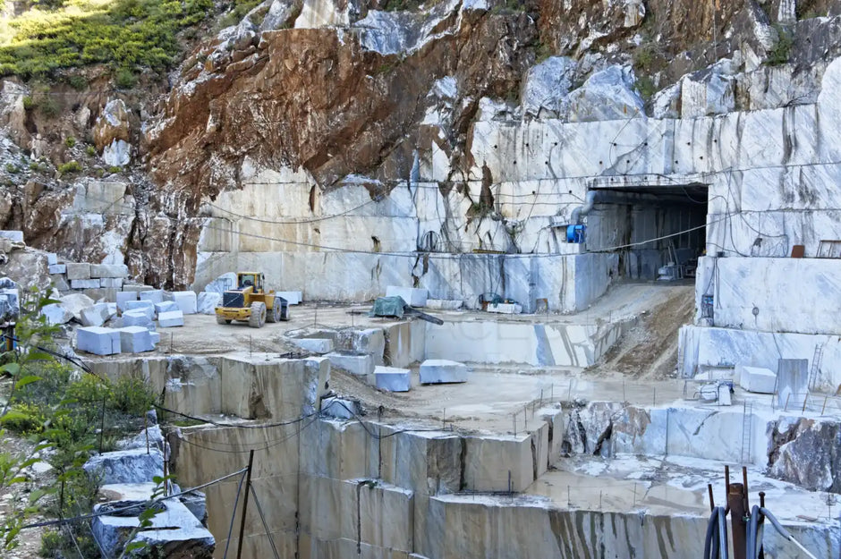 Marble quarry with large blocks of stone, a tunnel entrance, and construction equipment in a mountainous landscape.