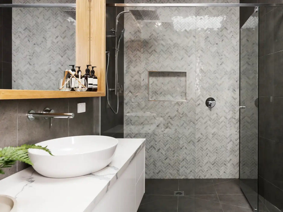 Modern bathroom with a walk-in glass shower, herringbone tile wall, vessel sink, and wooden shelf with toiletries.