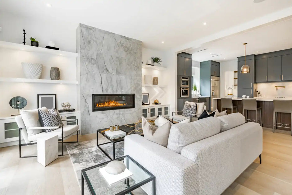 Modern living room with a gray sofa, marble fireplace, open shelving, and kitchen area in the background.