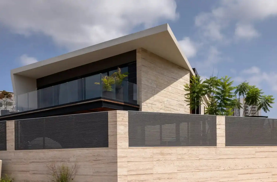 Modern two-story house with large glass windows, a sloped roof, and surrounded by a high concrete wall and greenery.