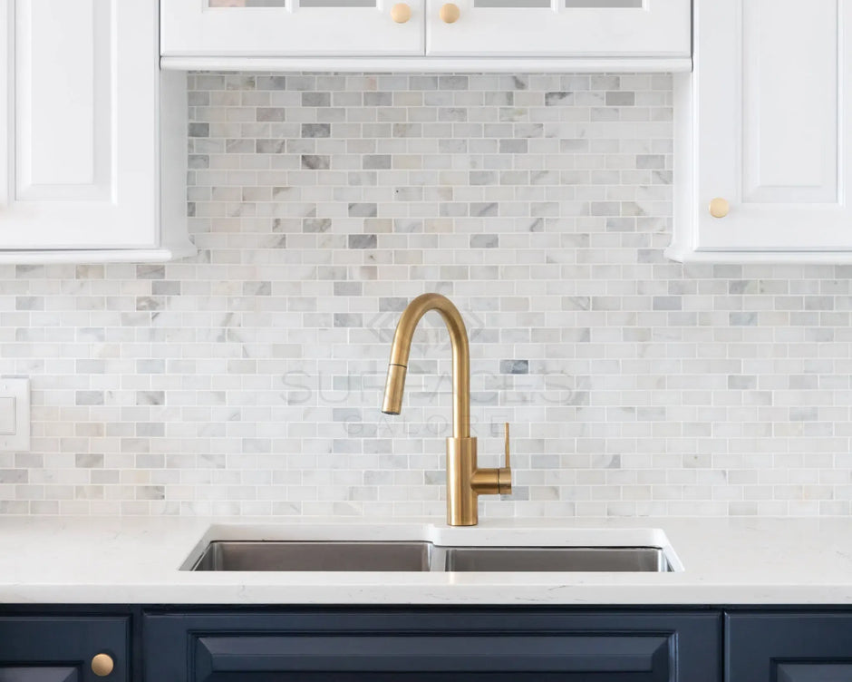 A kitchen sink with a gold faucet, marble tile backsplash, white cabinets, and a dark blue cabinet below the sink.