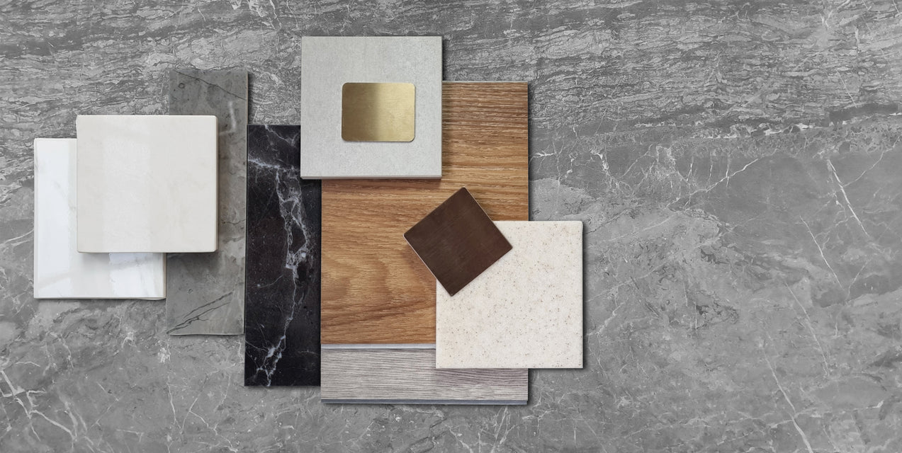 Various square and rectangular samples of wood, metal, marble, and stone arranged on a gray marbled surface.