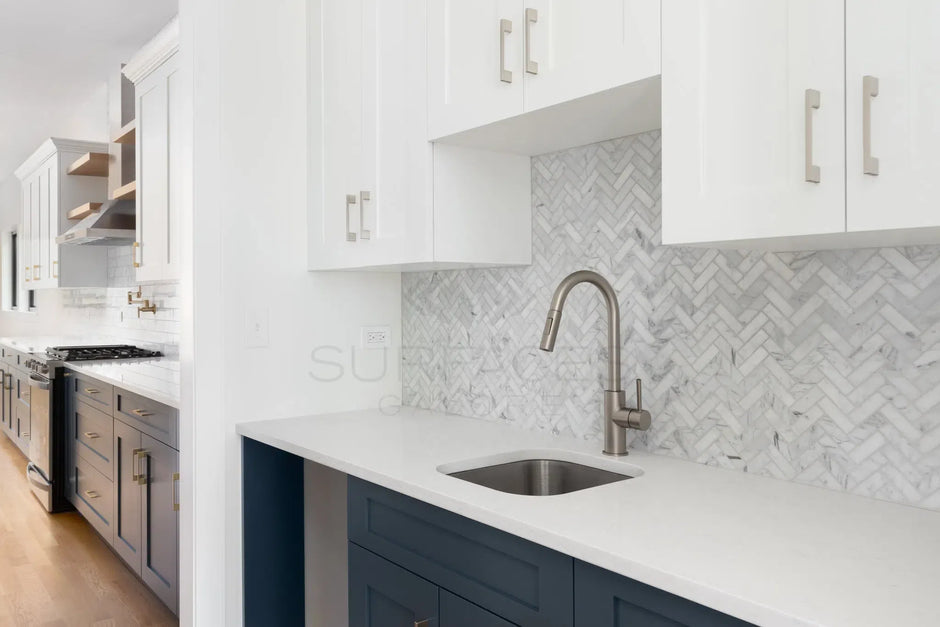 Modern kitchen with white and dark blue cabinetry, stainless steel fixtures, and herringbone tile backsplash.