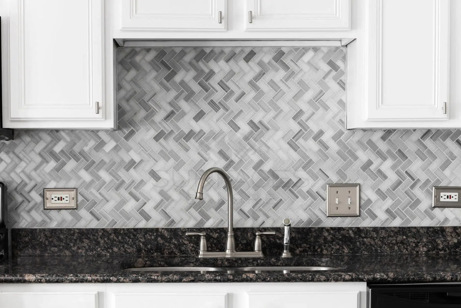 A kitchen with herringbone-patterned gray backsplash, white cabinets, and a stainless steel faucet and sink.