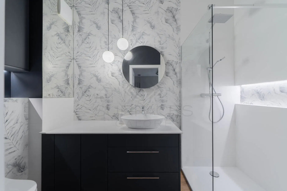 Modern bathroom with white vanity, round mirror, hanging lights, glass shower enclosure, and monochrome wallpaper.