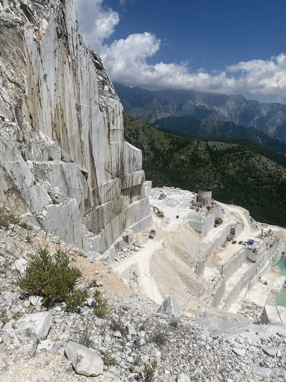 Expansive white marble quarry with green hills and mountains in the background under a blue sky with scattered clouds.