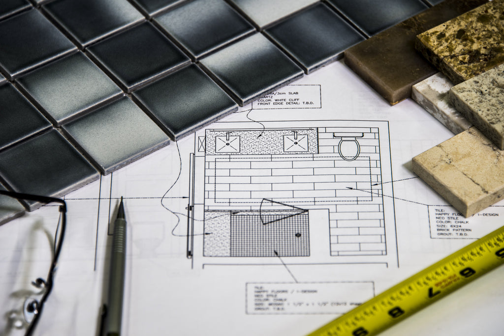 Architectural plan with tile samples, a pen, measuring tape, and glasses on a table.