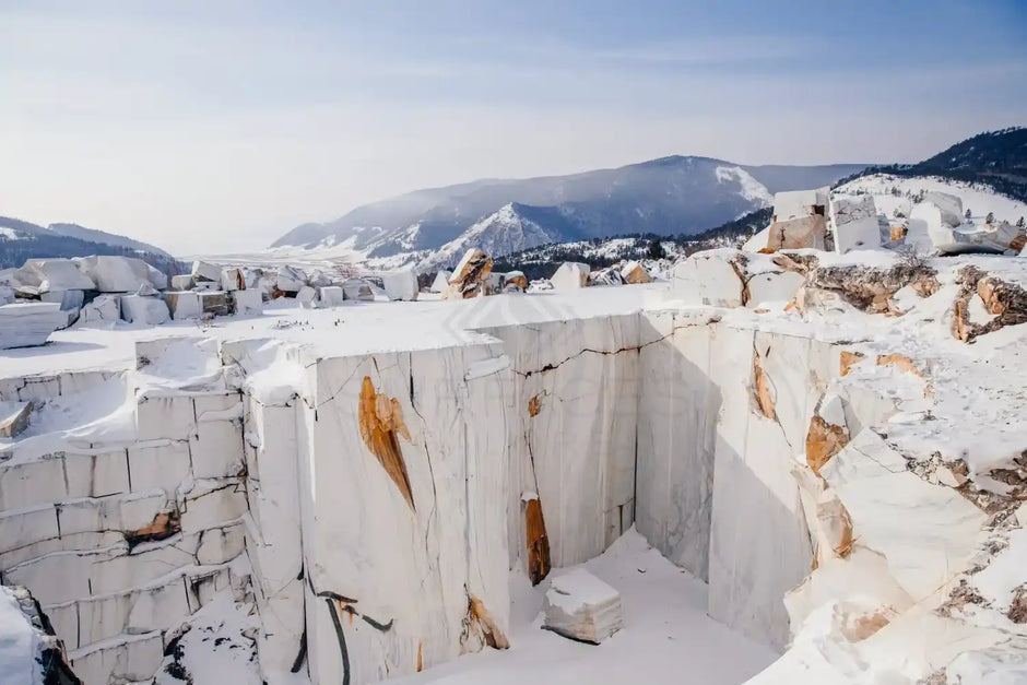 A snowy quarry with large white stone blocks, mountain range in the background under a clear blue sky.