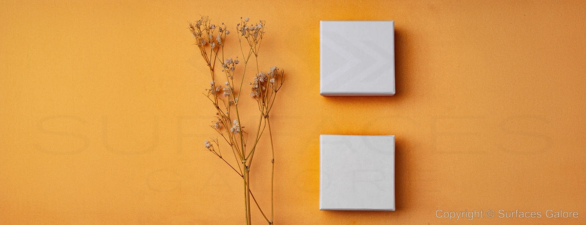 Two square white tiles are placed on an orange background with a small dried branch of delicate flowers on the left side. The text "Copyright © Surfaces Galore" is visible in the bottom right corner.