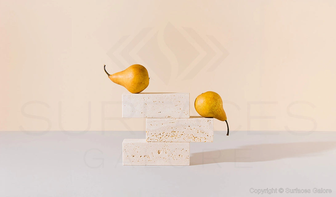 Two yellow pears are positioned artistically on a stack of three light beige rectangular stone blocks. The background is a soft, neutral shade, creating a minimalist and elegant aesthetic. The setup casts gentle shadows, enhancing the visual appeal.