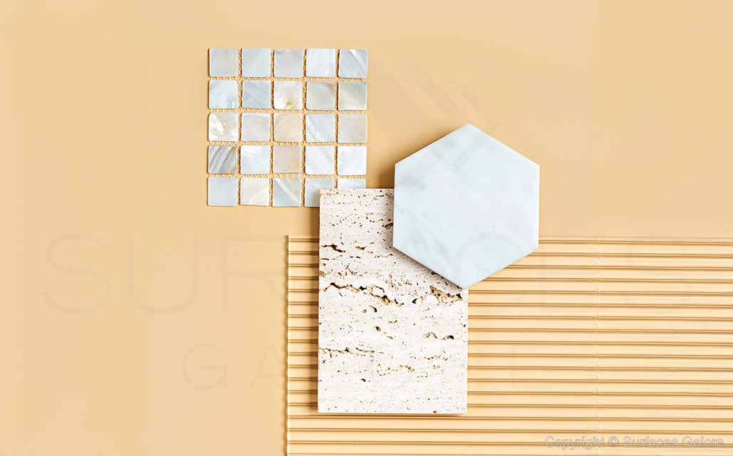 A flatlay of various tiles on a cream-colored background. The tiles include a square grid of small, pearl-like tiles, a hexagonal white marble tile, a rectangular beige stone tile with speckles, and a section of beige linear-textured tiles.