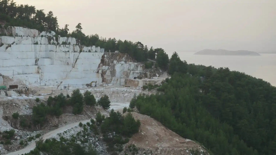 A coastal quarry with large white rock formations, surrounded by dense green trees and overlooking a distant island.