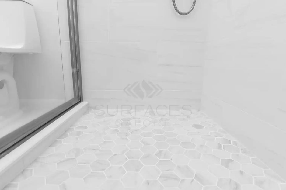 A shower with a glass door and a white hexagonal tile floor, featuring a toilet partially visible on the left.